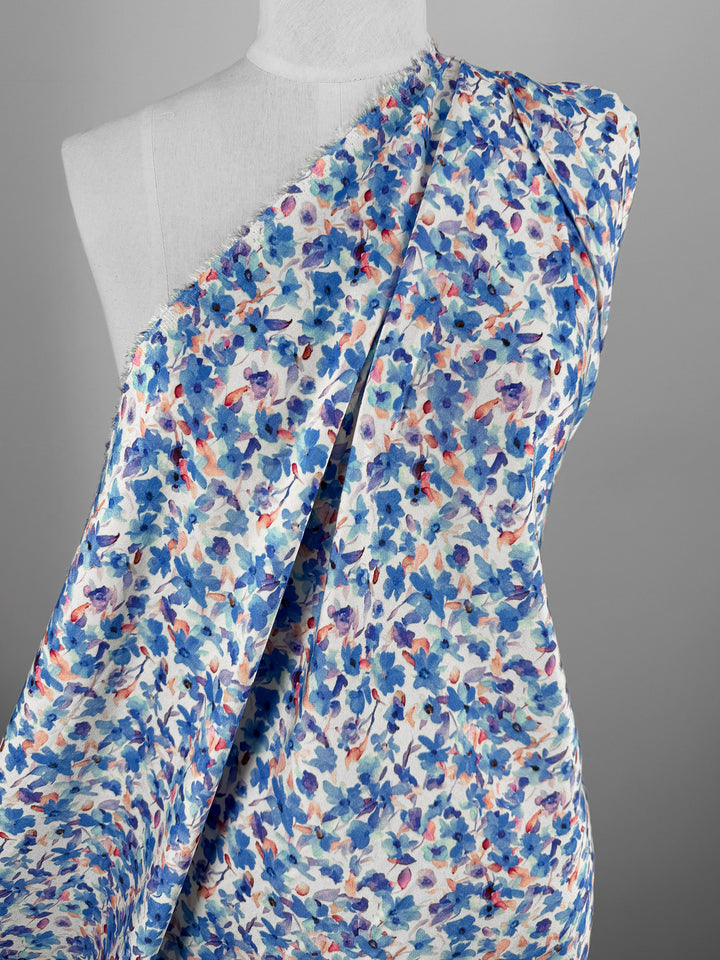 A white mannequin displays Super Cheap Fabrics’ Mercerised Rayon - Bluebell Drift (150cm), featuring a blue, purple, and orange floral print on white. The lightweight fabric is draped diagonally against a plain gray backdrop.