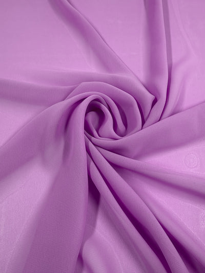 The Hi-Multi Chiffon - Violet Tulle (150cm) from Super Cheap Fabrics is displayed in soft, swirling folds of sheer light purple polyester, forming a spiral pattern at the center on a flat surface.