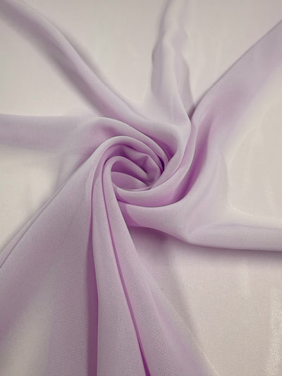 The Hi-Multi Chiffon - Orchid Petal - 150cm from Super Cheap Fabrics is elegantly swirled on a white surface, showcasing its light purple sheer texture, gentle folds, and delicate, airy beauty.