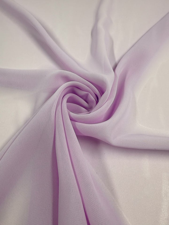 The Hi-Multi Chiffon - Orchid Petal - 150cm from Super Cheap Fabrics is elegantly swirled on a white surface, showcasing its light purple sheer texture, gentle folds, and delicate, airy beauty.