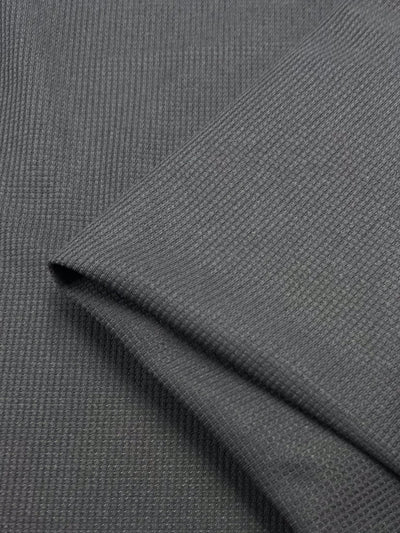 Close-up of Super Cheap Fabrics' Waffle Knit - Charcoal (170cm): dark grey, textured fabric with a subtle grid-like waffle pattern and a neatly folded edge overlapping the surface for a dimensional look.