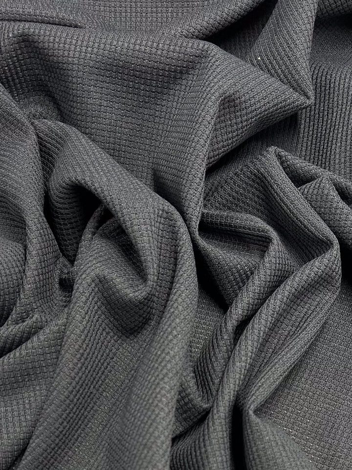 Super Cheap Fabrics’ Waffle Knit - Charcoal (170cm) features a dark gray, textured waffle weave with a three-dimensional look, loosely gathered and draped in soft folds.