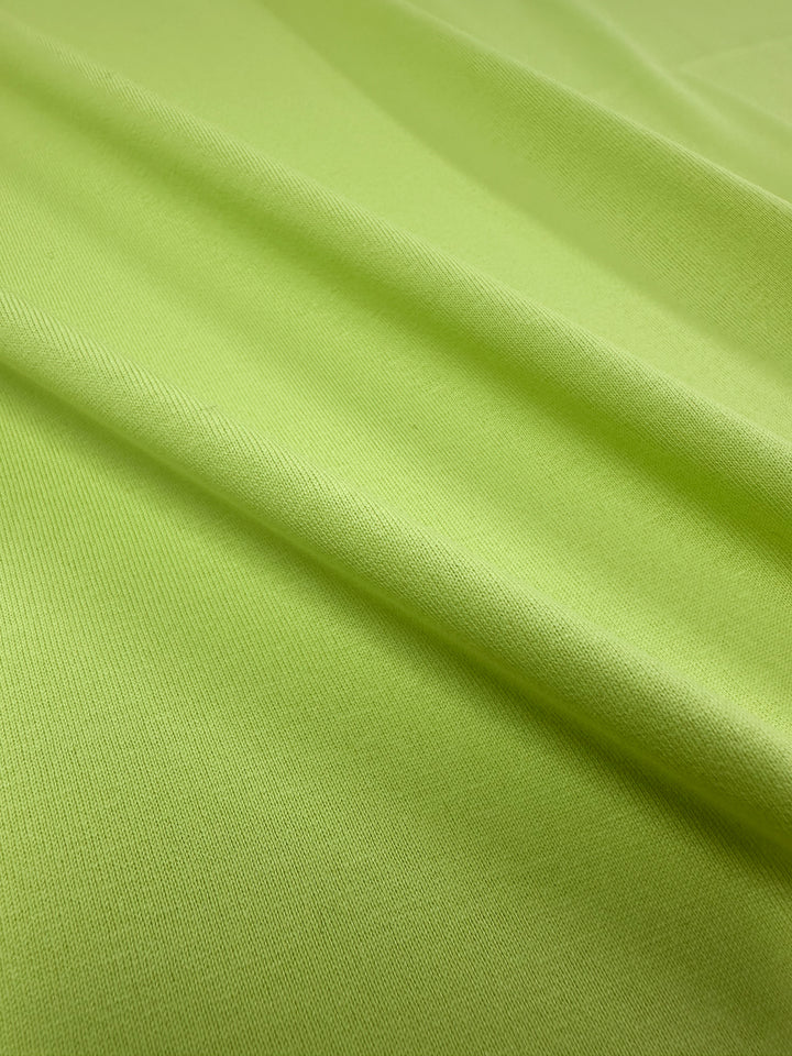 Close-up of Super Cheap Fabrics' Cotton Jersey in Daiquiri Green at 170cm wide, featuring a smooth, lime green appearance with soft folds and a subtle texture. This lightweight material is slightly shiny and resembles fine jersey fabric, with gentle shadows formed by the folds.