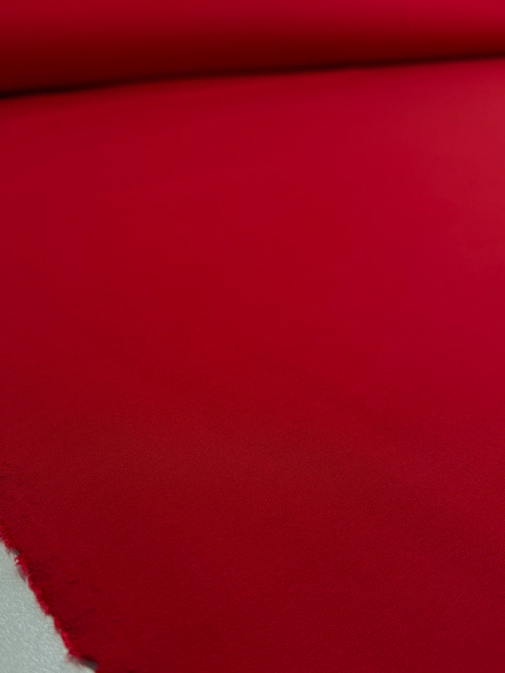 A close-up of Super Cheap Fabrics' Milano Ponte - True Red (160cm) shows the soft, smooth texture and slightly frayed edge as the bright red fabric is unrolled on a flat surface.