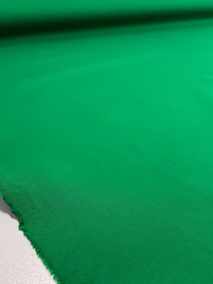 A close-up of Super Cheap Fabrics' Milano Ponte - Jolly Green (160cm) shows the smooth, heavy weight fabric laid flat with a slightly frayed edge, highlighting its even bright green color and fine texture.