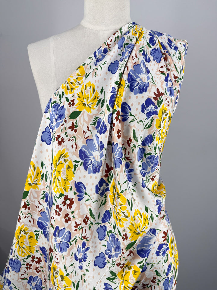 A dressmaker’s mannequin displays Super Cheap Fabrics’ Mercerised Rayon - Spring Bouquet 150cm, a lightweight, breathable fabric with a colorful yellow, blue, and burgundy floral print against a plain gray background.
