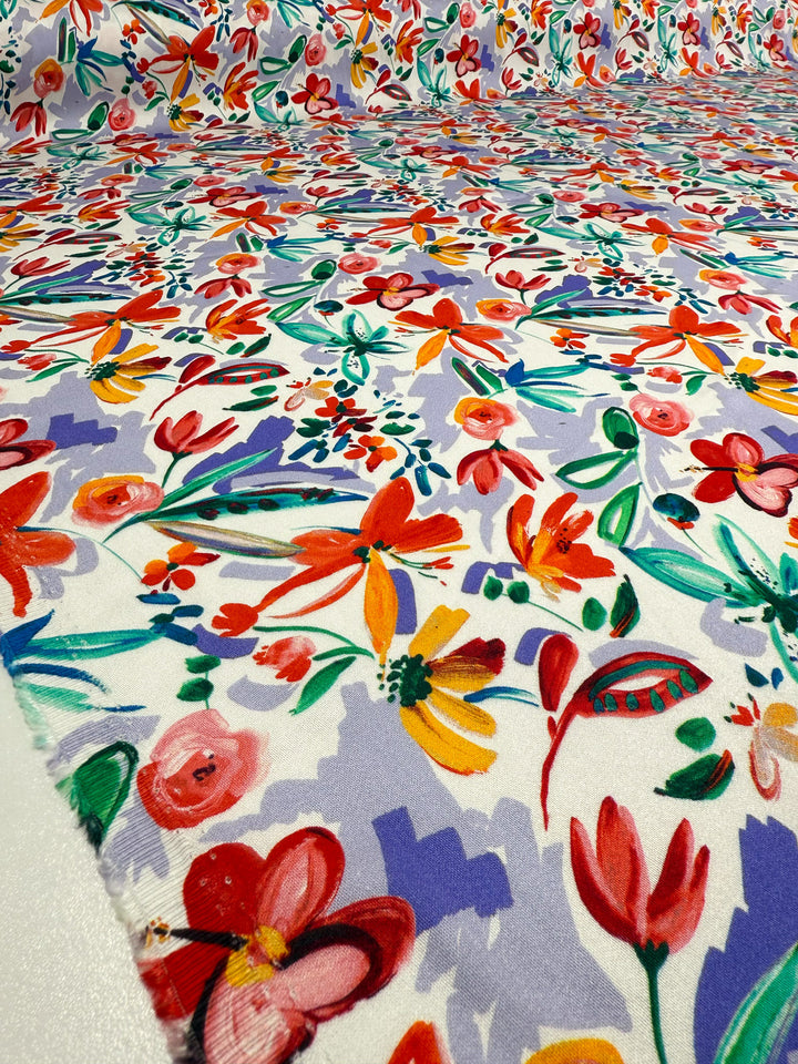 A close-up of Super Cheap Fabrics' Mercerised Rayon - Tropical Brushstroke, a lightweight 150cm fabric featuring vibrant red, yellow, pink, and orange flowers with green leaves on a white and light purple background.