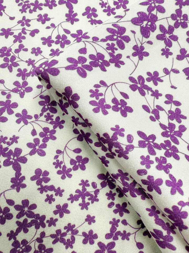 A close-up of Super Cheap Fabrics’ Mercerised Rayon - Violet Meadow (150cm) shows its lightweight, breathable fabric in a small violet floral and vine pattern, with a visible crease formed by a fold.