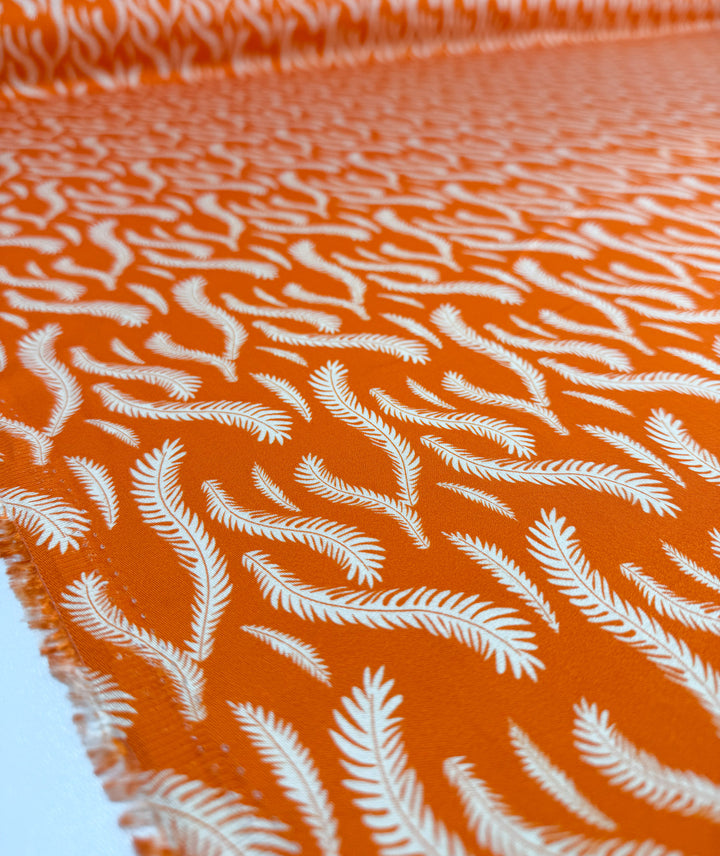 Close-up of Mercerised Rayon - Citrus Flow by Super Cheap Fabrics, a lightweight 150cm orange fabric with a white, fern-like repeat pattern and fluid drape, shown both flat and rolled for texture and vibrancy.