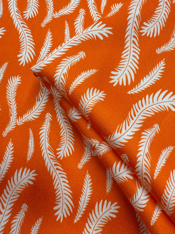 Super Cheap Fabrics’ Mercerised Rayon - Citrus Flow - 150cm is an orange fabric with a white leaf or feather pattern, showing overlapping sections that highlight the repeating design and its fluid drape.