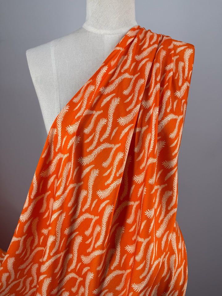 A dress form displays "Mercerised Rayon - Citrus Flow - 150cm" by Super Cheap Fabrics, a bright orange, lightweight rayon with a white fern-like leaf pattern, set against a plain gray background.