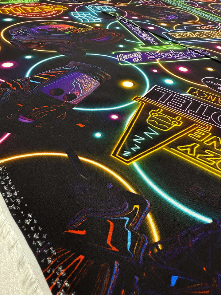 A close-up of Super Cheap Fabrics’ Designer Rayon - After Dark (140cm) shows vibrant neon-style prints with illustrated signs, shapes, and glowing outlines in various colors on a black background.