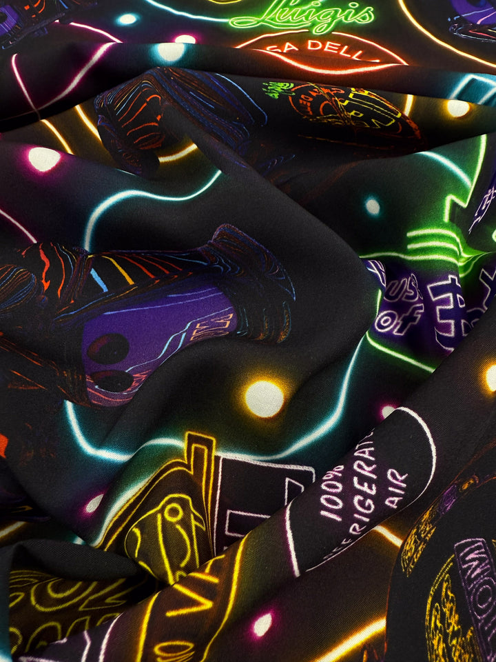 A close-up of Super Cheap Fabrics’ Designer Rayon - After Dark (140cm), showing vibrant neon car, text, and shape prints in green, purple, yellow, and blue on black. Fabric is slightly folded.