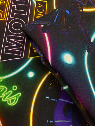 Close-up of Designer Rayon - After Dark - 140cm by Super Cheap Fabrics, showcasing neon "MOTEL" and "VACANCY" prints with glowing circles and abstract shapes in bold purples, blues, yellows, and greens.