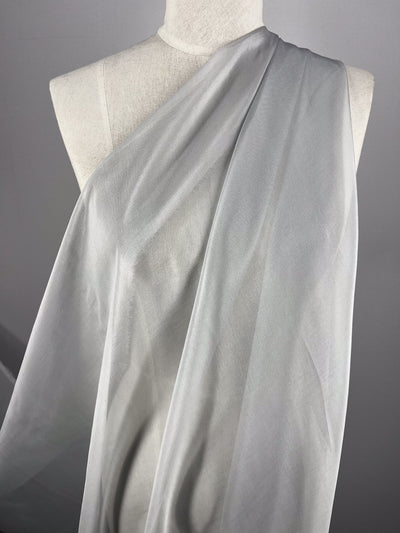 A white mannequin displays the subtle metallic shimmer of Super Cheap Fabrics' Silk Mousseline in Moonbeam. This sheer, light gray fabric cascades diagonally, creating soft, flowing lines against a simple gray backdrop. Fabric width: 140cm.