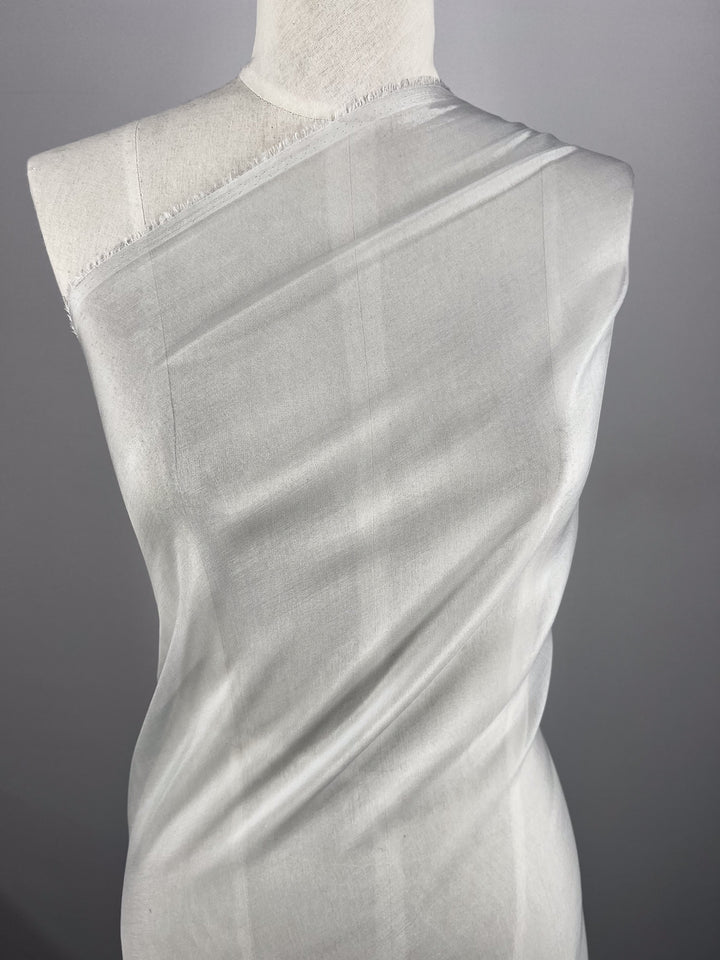 A white mannequin displays "Silk Mousseline - Moonbeam - 140cm" by Super Cheap Fabrics, draped in sheer moonbeam-colored fabric against a gray background, with the silk asymmetrically wrapped across its chest and shoulder to highlight its texture and translucence.