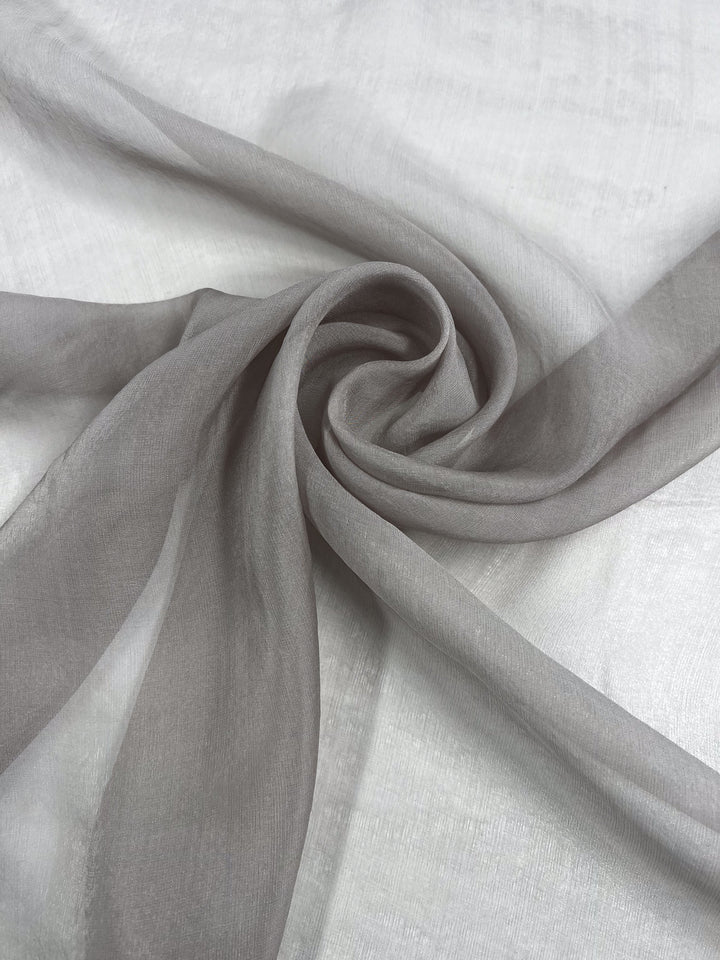 Super Cheap Fabrics' Silk Mousseline - Marble - 140cm is a sheer, light gray silk with a subtle metallic shimmer, delicately arranged in soft folds to showcase its airy, extra lightweight texture on a flat white surface.