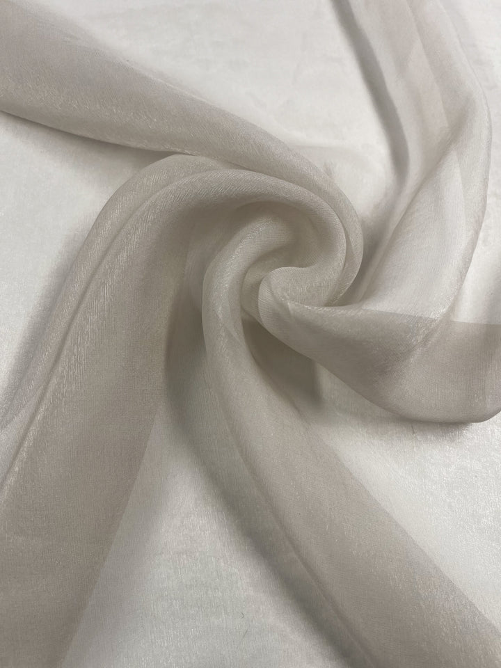 The Silk Mousseline - Oyster from Super Cheap Fabrics, measuring 140cm, displays sheer light gray fabric in artistic swirling folds on a flat surface, showcasing its delicate texture and metallic shimmer.