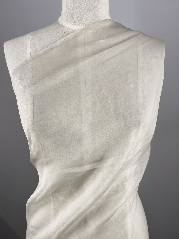 A mannequin showcases the Silk Mousseline - Oyster - 140cm by Super Cheap Fabrics, featuring an extra light, sheer material with a delicate metallic shimmer draped over its shoulder and back. The fabric's translucency and subtle texture stand out elegantly against the neutral background.