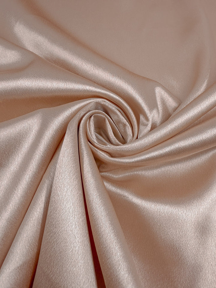 A close-up of Super Cheap Fabrics' Satin Back Crepe in Cream Tan (150cm), showing its smooth, shiny texture arranged in gentle folds with a spiral swirl at the center.