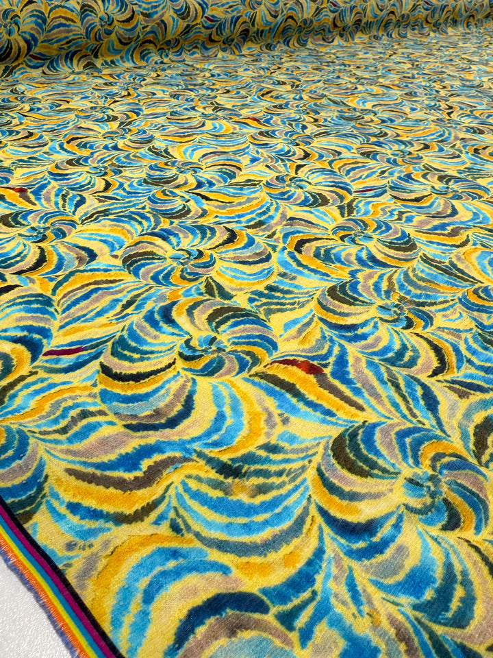 Super Cheap Fabrics’ Textured Designer Cotton - Spiral Sun (145cm) features vibrant yellow, blue, teal, and brown spiral wave designs with a colorful striped selvage edge.