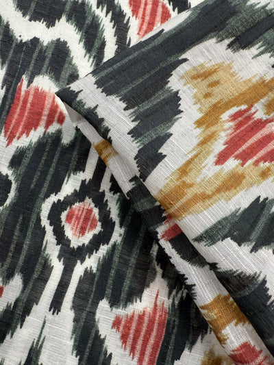 Close-up of Super Cheap Fabrics' Textured Designer Cotton - Spice Loom (150cm), featuring an Ikat pattern in black, white, red, and mustard yellow with bold shapes and visible texture. Lightweight cotton is shown folded over another piece.