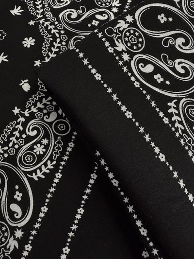 Close-up of Designer Rayon - Bandana Noir (148cm) by Super Cheap Fabrics: black lightweight rayon with a white paisley and floral bandana pattern, featuring intricate swirls, flowers, and dotted lines.