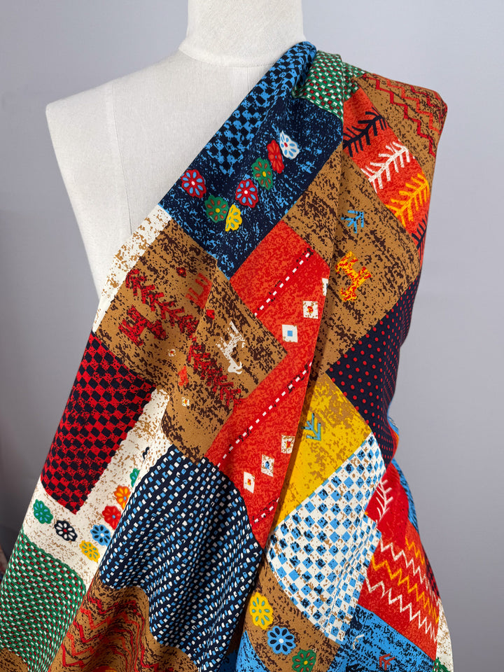 Super Cheap Fabrics’ Designer Rayon - Andes Patchwork (147cm) is draped on a mannequin, showcasing its vibrant geometric, floral, dot, and zigzag design in red, blue, yellow, brown, and white.