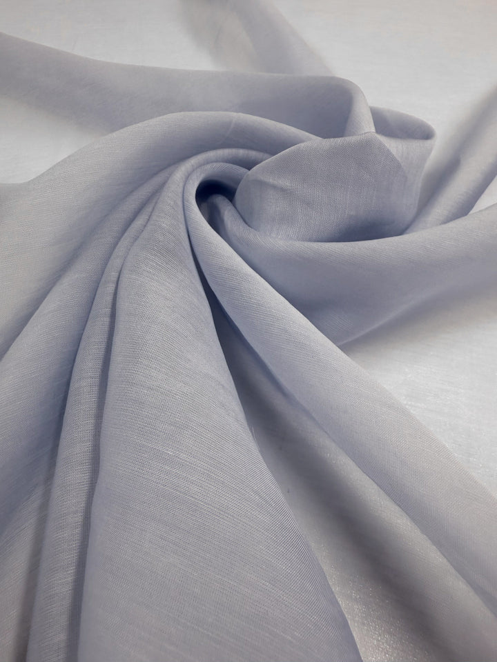 A close-up of Super Cheap Fabrics' Designer Voile - Skywriting (150cm), a light gray, lightweight fabric loosely spiraled on a smooth surface, highlights its soft texture and semi-transparent quality.