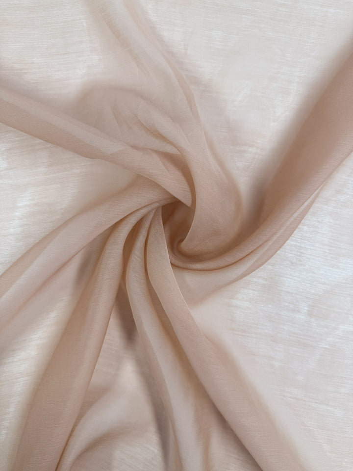 Designer Voile - Apple Blossom - 150cm by Super Cheap Fabrics features sheer, light beige fabric loosely gathered in swirls on a flat surface, creating soft folds and an airy, delicate texture.