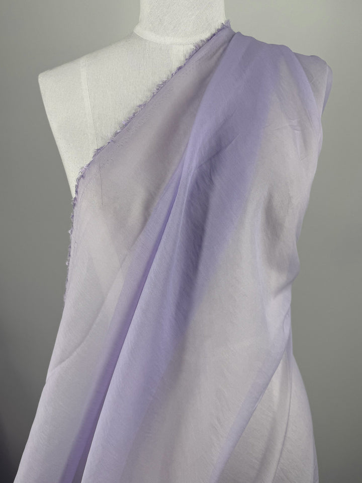 A mannequin displays Super Cheap Fabrics’ Designer Voile - Pastel Lilac - 150cm, draped diagonally to reveal one shoulder. The sheer, lightweight cotton voile creates airy elegance against a plain neutral background.
