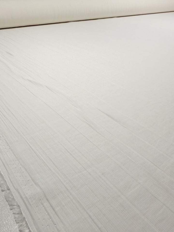 A large roll of Super Cheap Fabrics’ Textured Designer Voile in Ivory (155cm wide) is unrolled on a flat surface, displaying its smooth, slightly textured cotton with subtle lines and frayed edges.