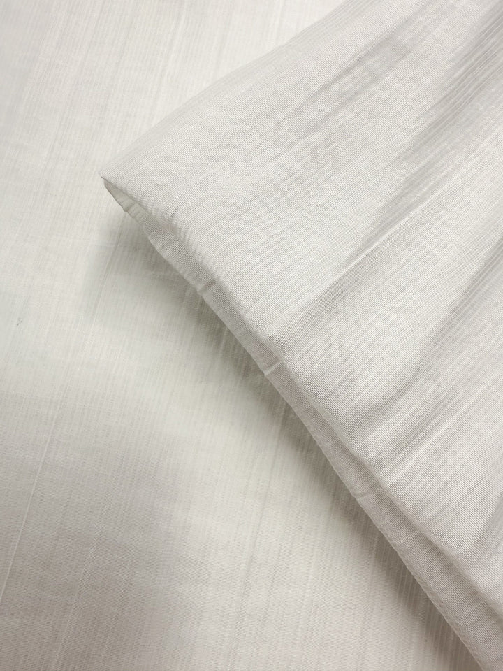 A close-up of Super Cheap Fabrics' Textured Designer Voile - Ivory - 155cm, showcasing its light color, soft texture, and slightly wrinkled look as it rests on matching fabric.