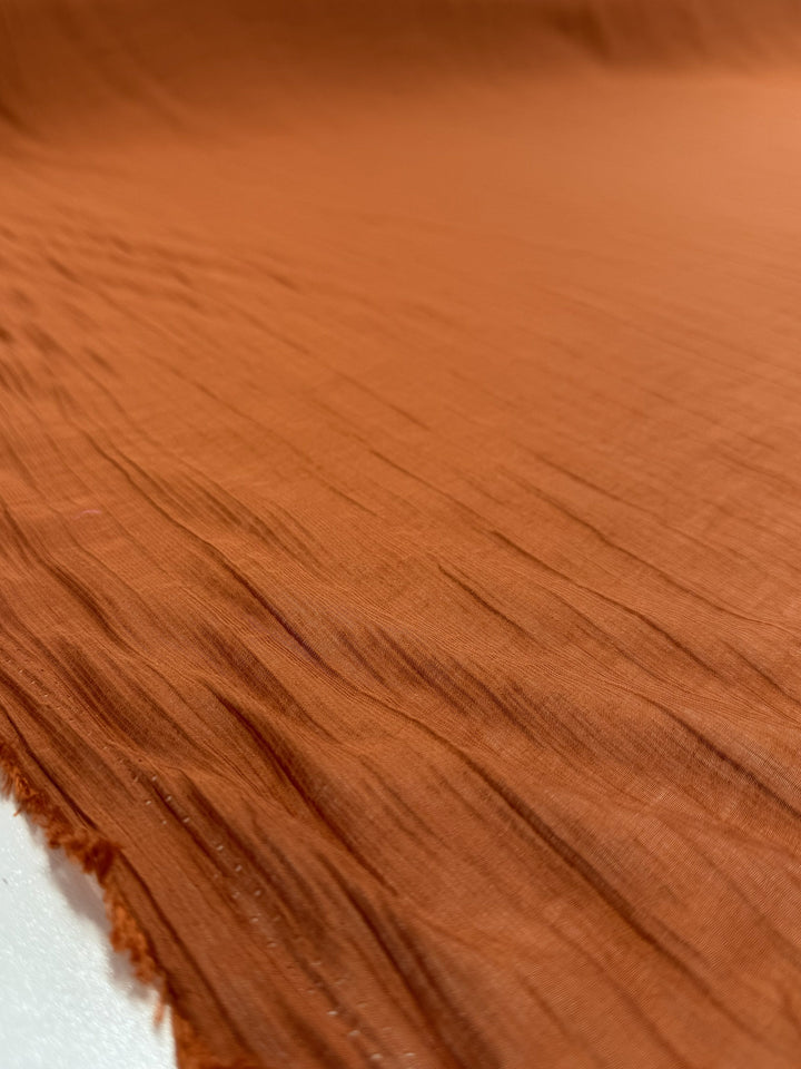 A close-up of Super Cheap Fabrics' Textured Designer Voile - Rust - 155cm shows a lightweight, rust-colored fabric with fine lines, natural irregularities, and a slightly crinkled surface. Soft texture and raw edge are visible in the foreground.