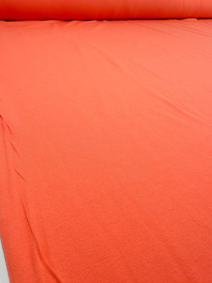A large roll of Super Cheap Fabrics' Cotton Lycra - Fusion Coral - 170cm lies partially unrolled, revealing its smooth texture. The medium-heavy weight fabric displays a vibrant hue across the entire visible area, creating a vivid and uniform appearance.