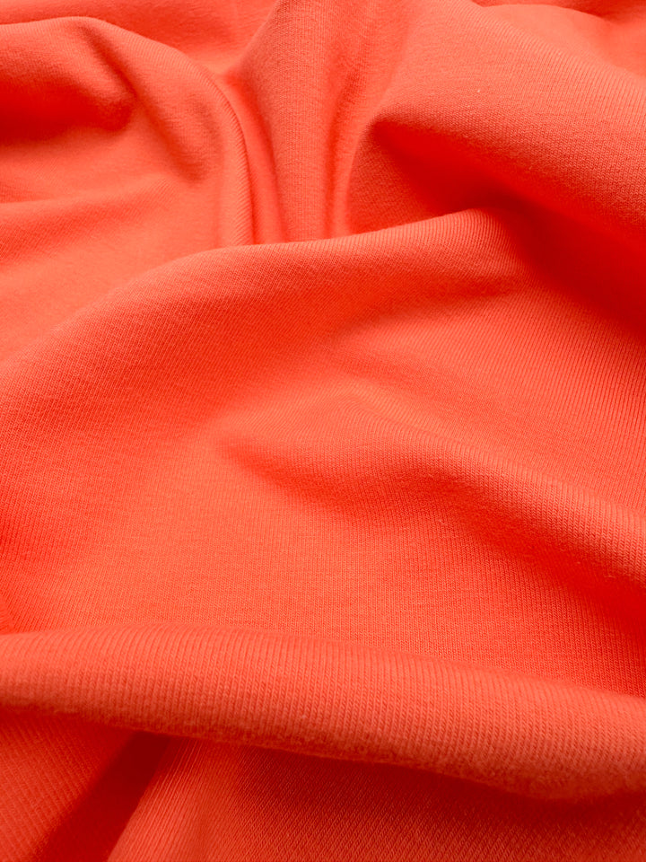 The Cotton Lycra - Fusion Coral by Super Cheap Fabrics presents a close-up view of its bright orange, ribbed texture. This fabric drapes softly with gentle waves and folds, showcasing its smooth and stretchy quality across 170cm.