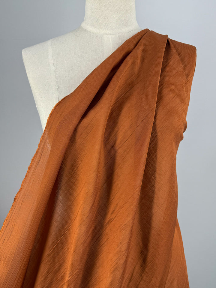 A mannequin showcases the Super Cheap Fabrics Textured Designer Voile - Rust - 155cm, a lightweight cotton-nylon blend with a subtle sheen, displayed against a plain light gray background.