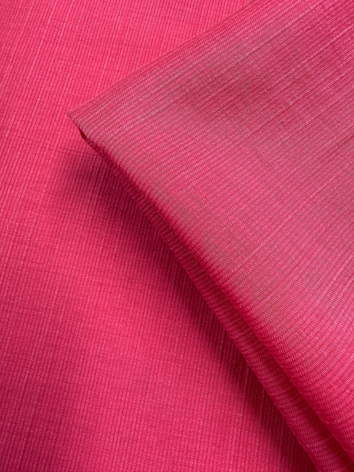 A close-up of Super Cheap Fabrics’ Textured Designer Voile - Raspberry Sorbet (155cm) shows two pieces—one flat, one folded—emphasizing the lightweight ribbed texture and intricate weave of this vibrant fabric.