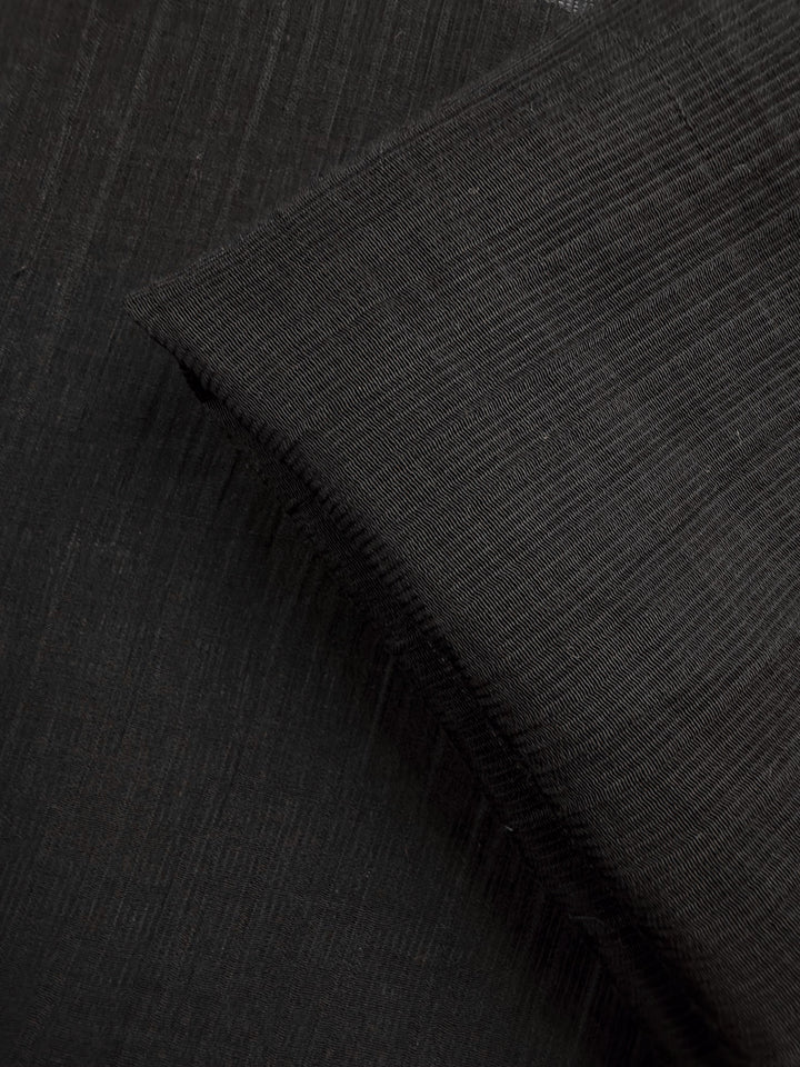 Close-up of Super Cheap Fabrics' Textured Designer Voile - Black (155cm), featuring fine horizontal and vertical lines with a folded corner that highlights its subtle weave—ideal for creating sheer summer dresses.