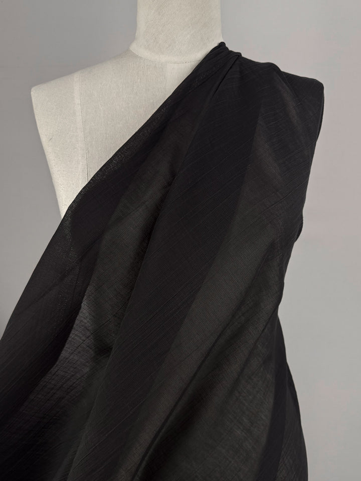 A torso mannequin displays Super Cheap Fabrics' Textured Designer Voile - Black - 155cm. The sheer, lightly textured fabric drapes over one shoulder and flows diagonally against a neutral background, highlighting its elegant, lightweight quality.
