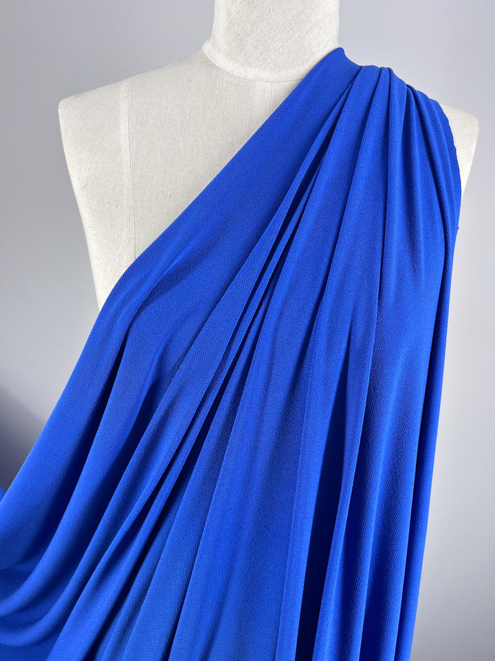 The Super Cheap Fabrics' Crepe Lycra in Sapphire (148cm) elegantly drapes over a mannequin with its one-shoulder design. The medium-weight fabric forms soft pleats, highlighting its smooth texture and vibrant color against a neutral light gray background.