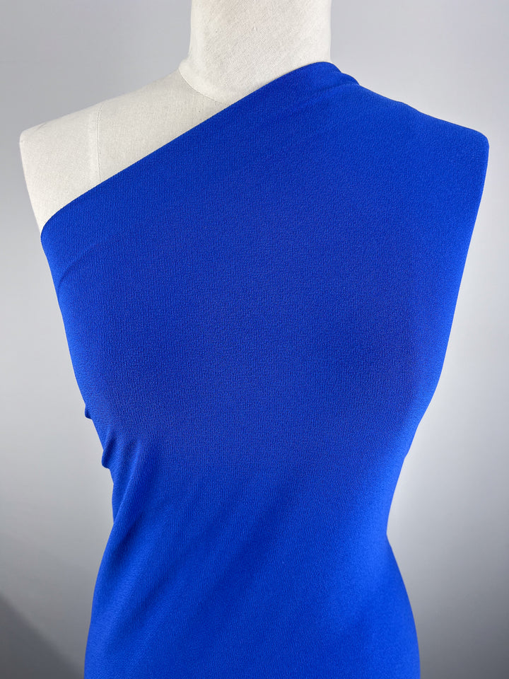 A mannequin showcases Super Cheap Fabrics' Crepe Lycra in vibrant sapphire against a gray backdrop; the material's smooth texture and medium-weight jersey contribute to its simple, elegant appeal.