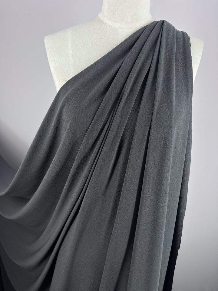 A white mannequin is elegantly draped in Super Cheap Fabrics' Crepe Lycra - Squid, 148cm, with a graceful gathered effect on one shoulder. The medium-weight jersey highlights its texture and softness in flowing gray.