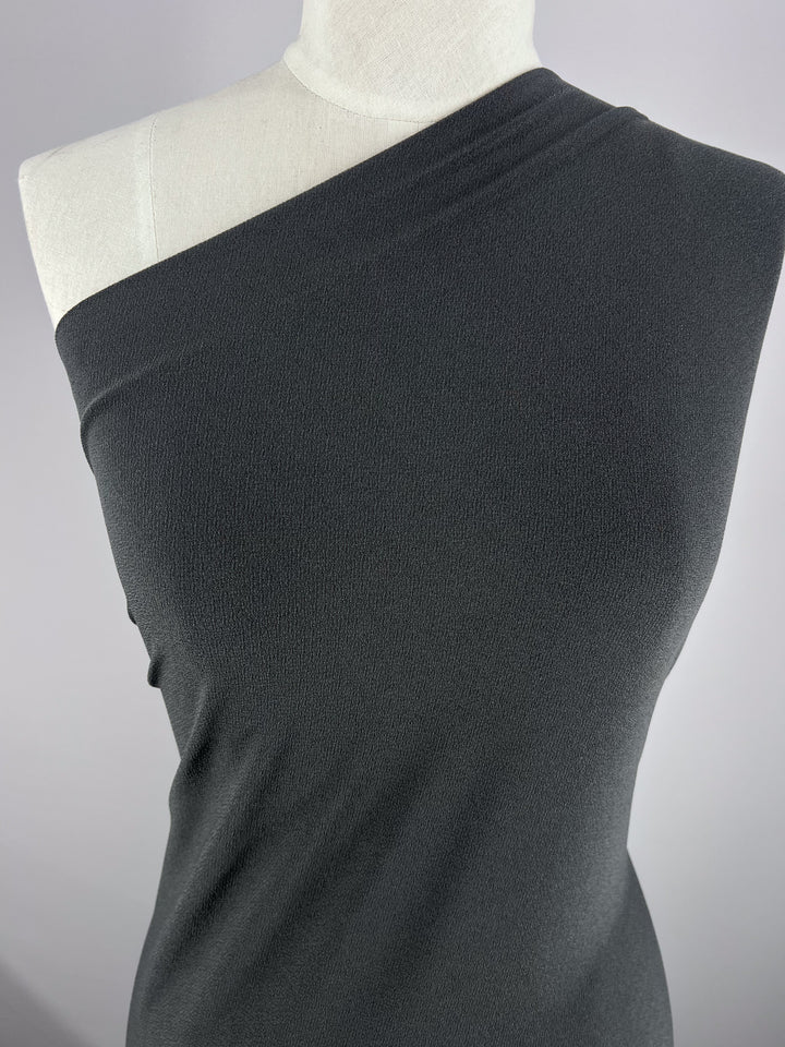 A mannequin displays a dark gray, one-shoulder, sleeveless top crafted from Super Cheap Fabrics' Crepe Lycra - Squid, 148cm. The polyester blend is soft and slightly textured against a plain light gray background.