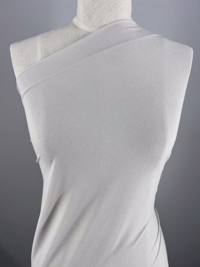 A mannequin displays a sleeveless, asymmetrical silver dress made from Crepe Lycra by Super Cheap Fabrics. The medium-weight material presents a subtle texture, with an elegant neckline crossing diagonally from shoulder to the opposite underarm against a plain gray backdrop.