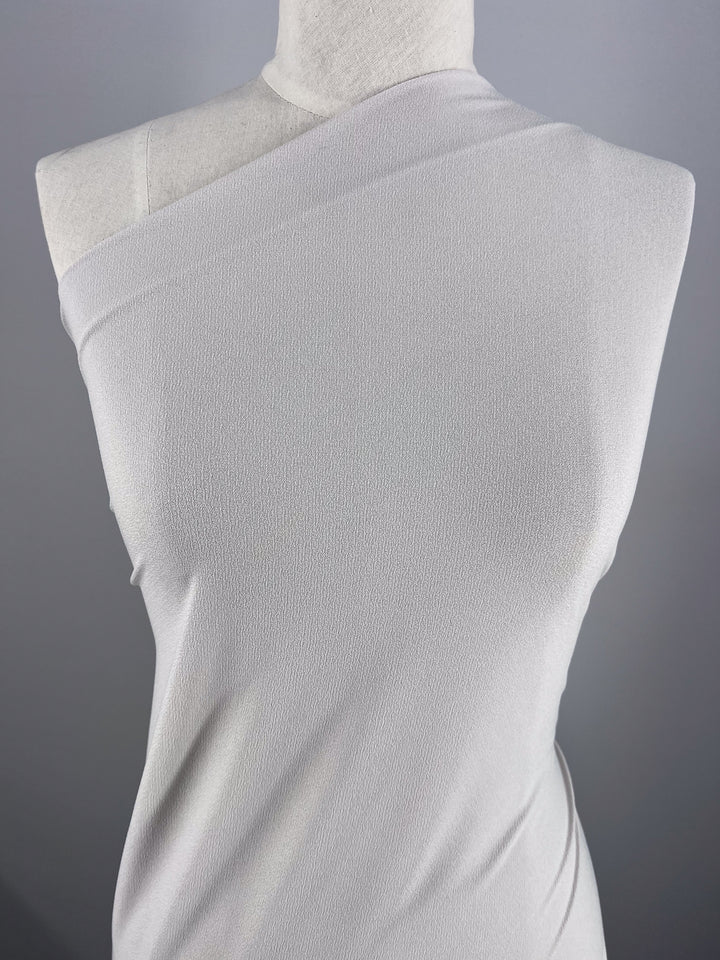 A mannequin displays a sleeveless, asymmetrical silver dress made from Crepe Lycra by Super Cheap Fabrics. The medium-weight material presents a subtle texture, with an elegant neckline crossing diagonally from shoulder to the opposite underarm against a plain gray backdrop.