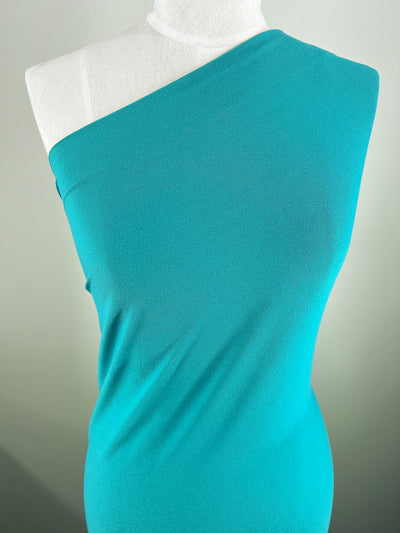A turquoise, one-shoulder dress in "Crepe Lycra - Jade - 148cm" from Super Cheap Fabrics is beautifully showcased on a dress form against a light gray background. The medium-weight jersey blend highlights its fitted silhouette, radiating simple sophistication.