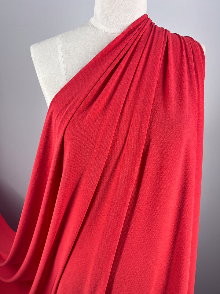 A mannequin draped in Super Cheap Fabrics' Crepe Lycra - Tangelo showcases vibrant coral pink fabric, gathered at the shoulder and elegantly cascading. The material's texture and pleats are highlighted against a neutral gray background.