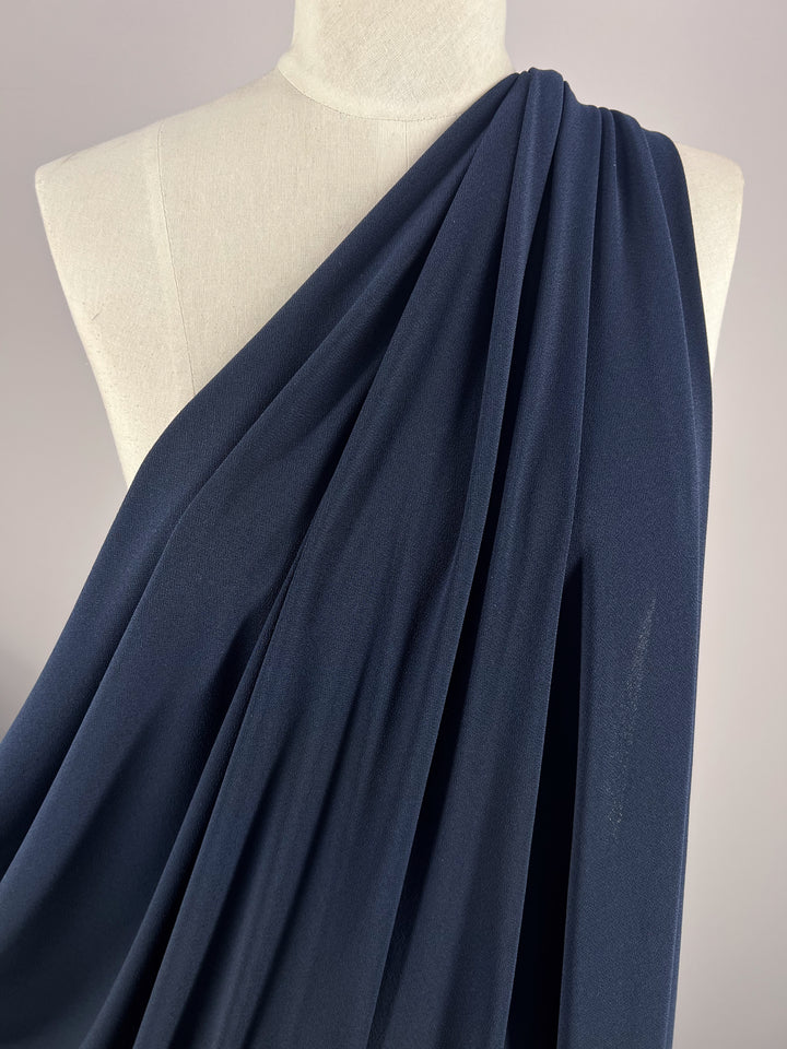 A mannequin showcases the Crepe Lycra - Ink - 148cm by Super Cheap Fabrics, draped over one shoulder with cascading folds. The plain light gray background emphasizes the fabric's rich texture and dark navy color.