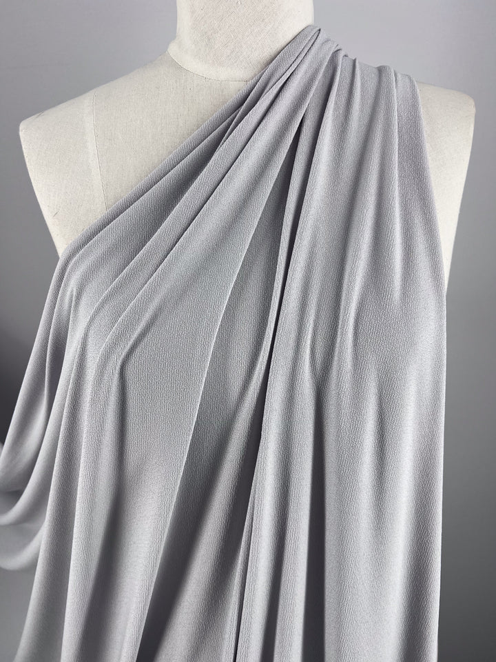 A close-up of a mannequin draped in Super Cheap Fabrics' Crepe Lycra in Smoke (148cm), showcasing elegant folds cascading over one shoulder, set against a plain gray background.
